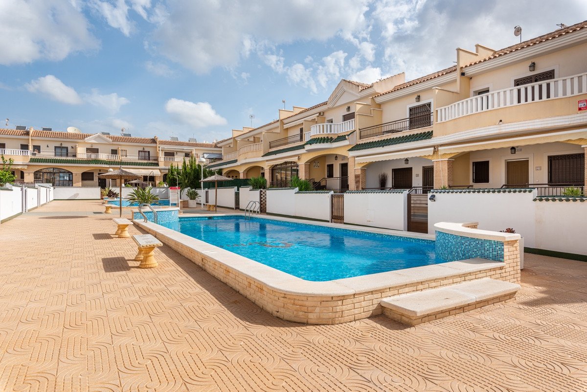 A bright outdoor shot of a swimming pool and adjacent terraced houses under a blue sky with clouds.