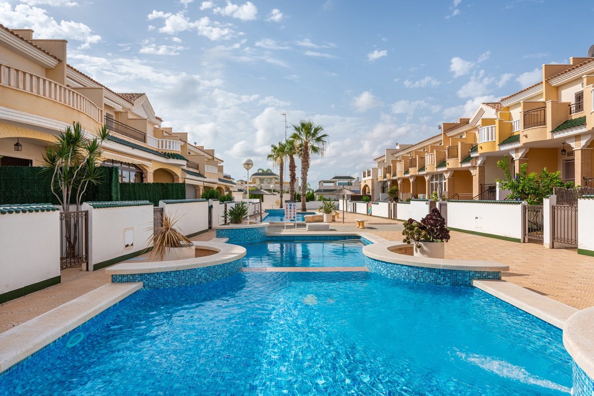 Luxurious swimming pool area with palm trees and modern townhouses under a blue sky with clouds.