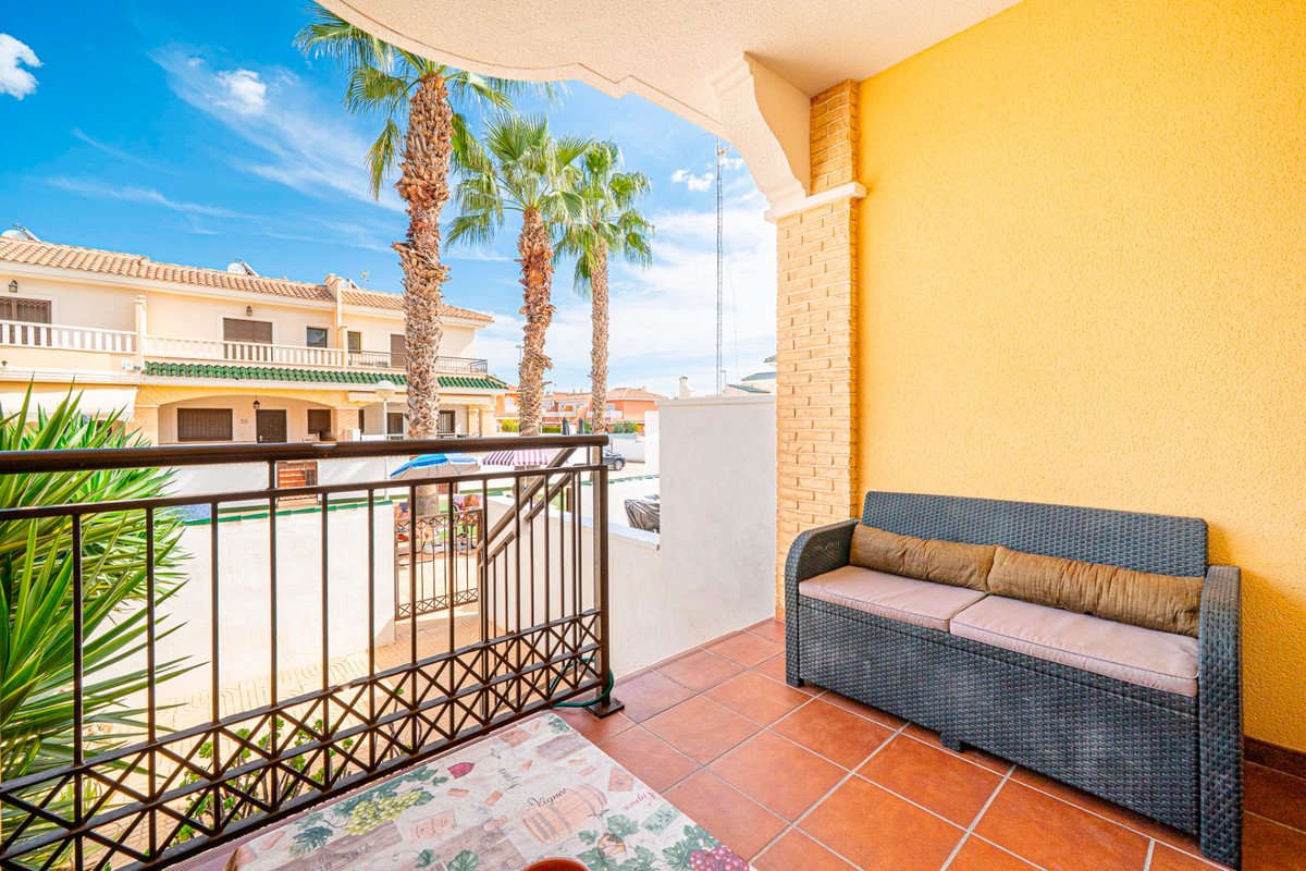A sunny balcony overlooking palm trees, a pool, and residential buildings. Features a black rattan sofa.