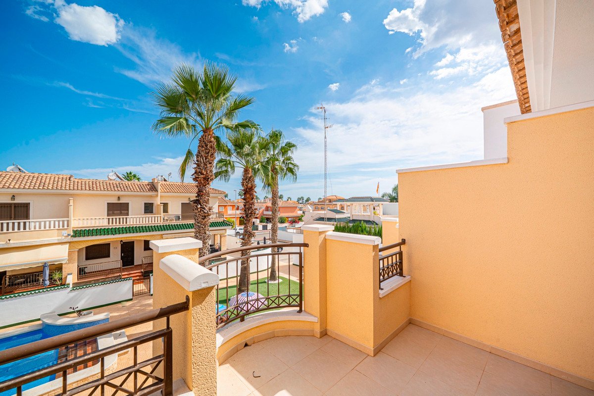 Balcony overlooking a swimming pool and palm trees with residential buildings in the background under a blue sky.