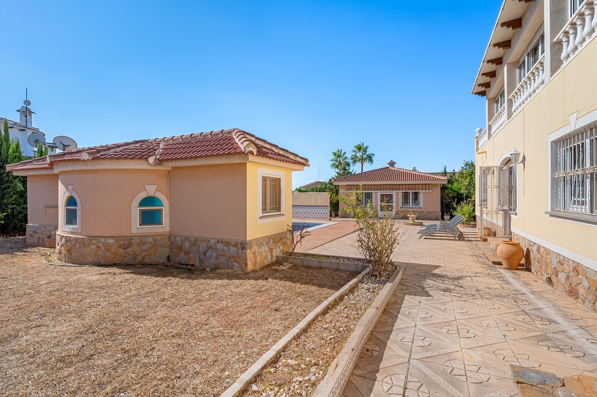 View of a villa with a pool, patio, and a separate guest house under a clear blue sky.