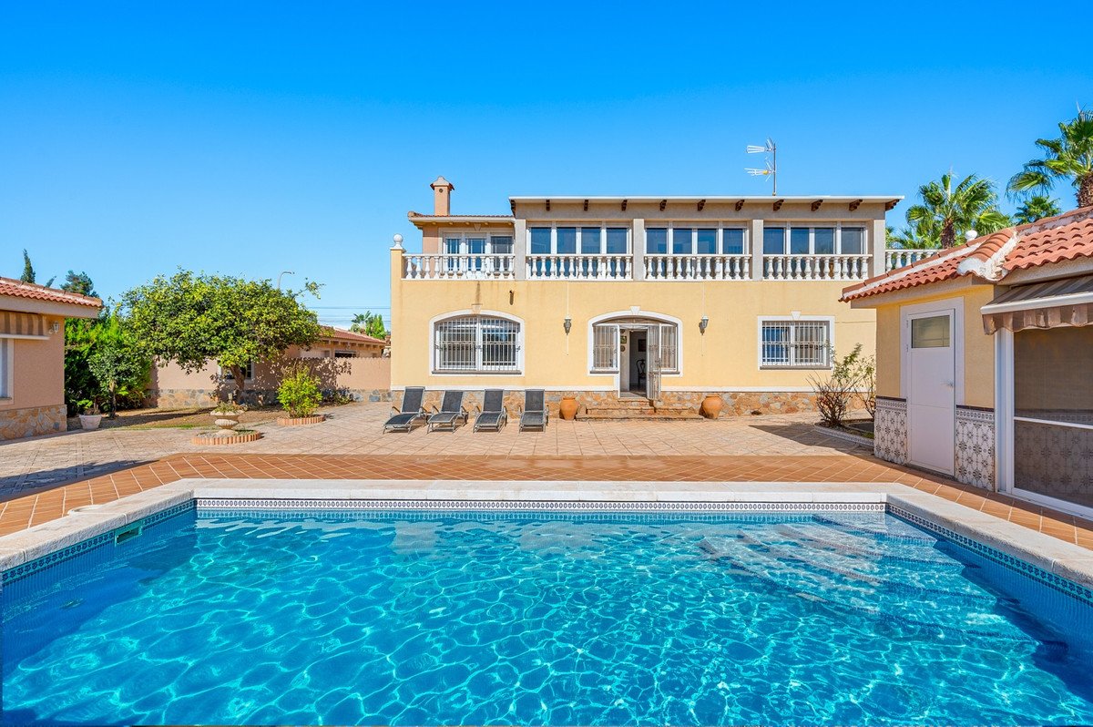 Yellow villa with a large blue swimming pool in the foreground, lounge chairs on the patio, and lush greenery under a clear blue sky.