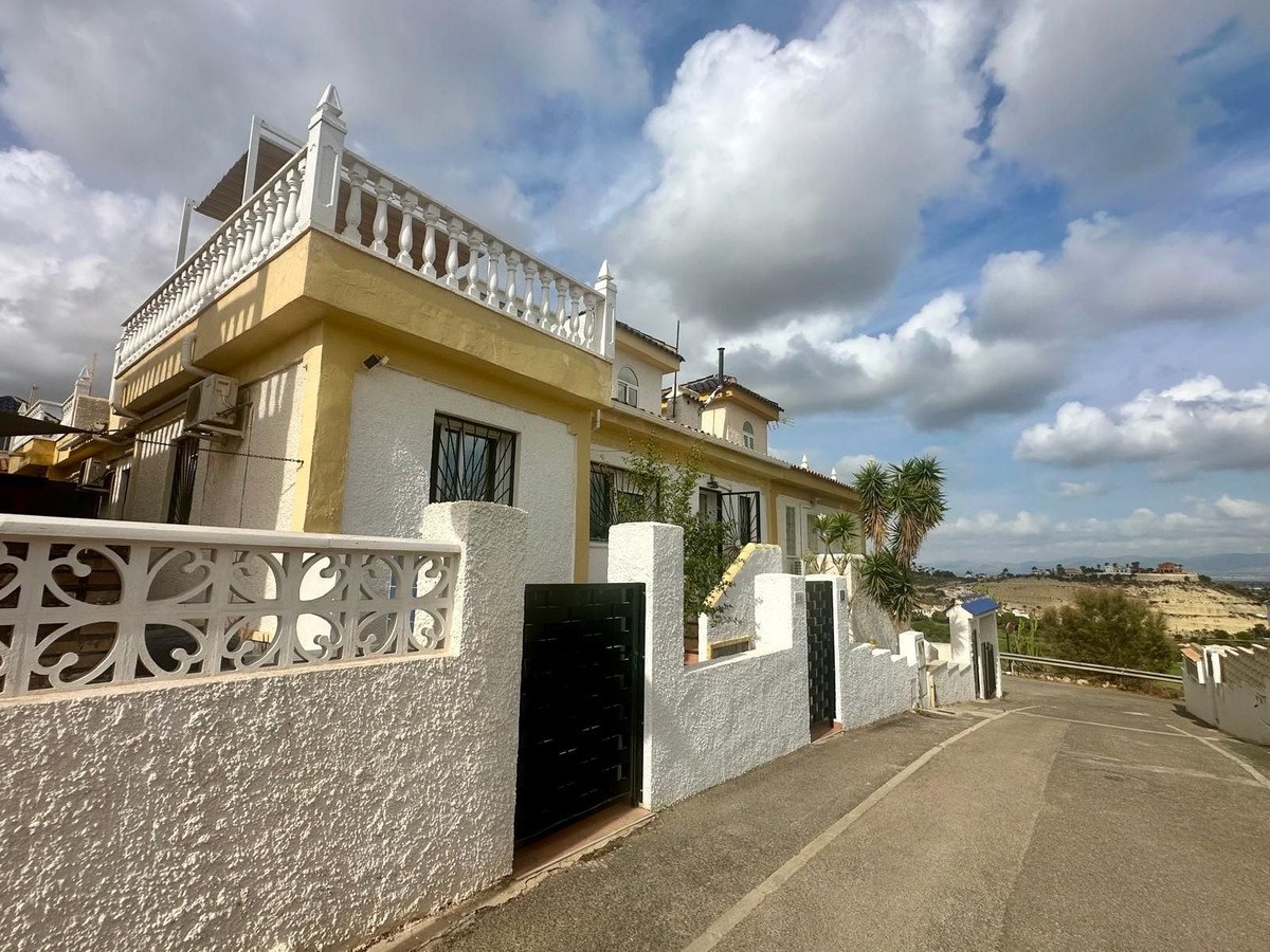 A yellow and white villa with a decorative white fence and a view of distant hills under a cloudy sky.
