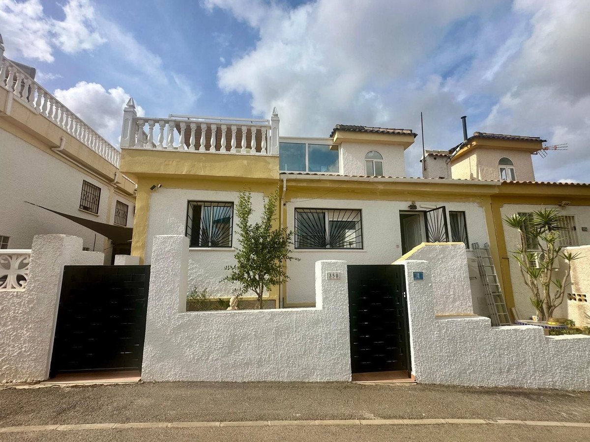 Exterior view of a white and yellow townhouse with black gates, a balcony, and a small tree in front under a cloudy sky.