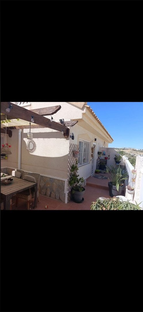 Terrace with a wooden dining table, potted plants, and a pergola overlooking a hillside view under a clear blue sky.