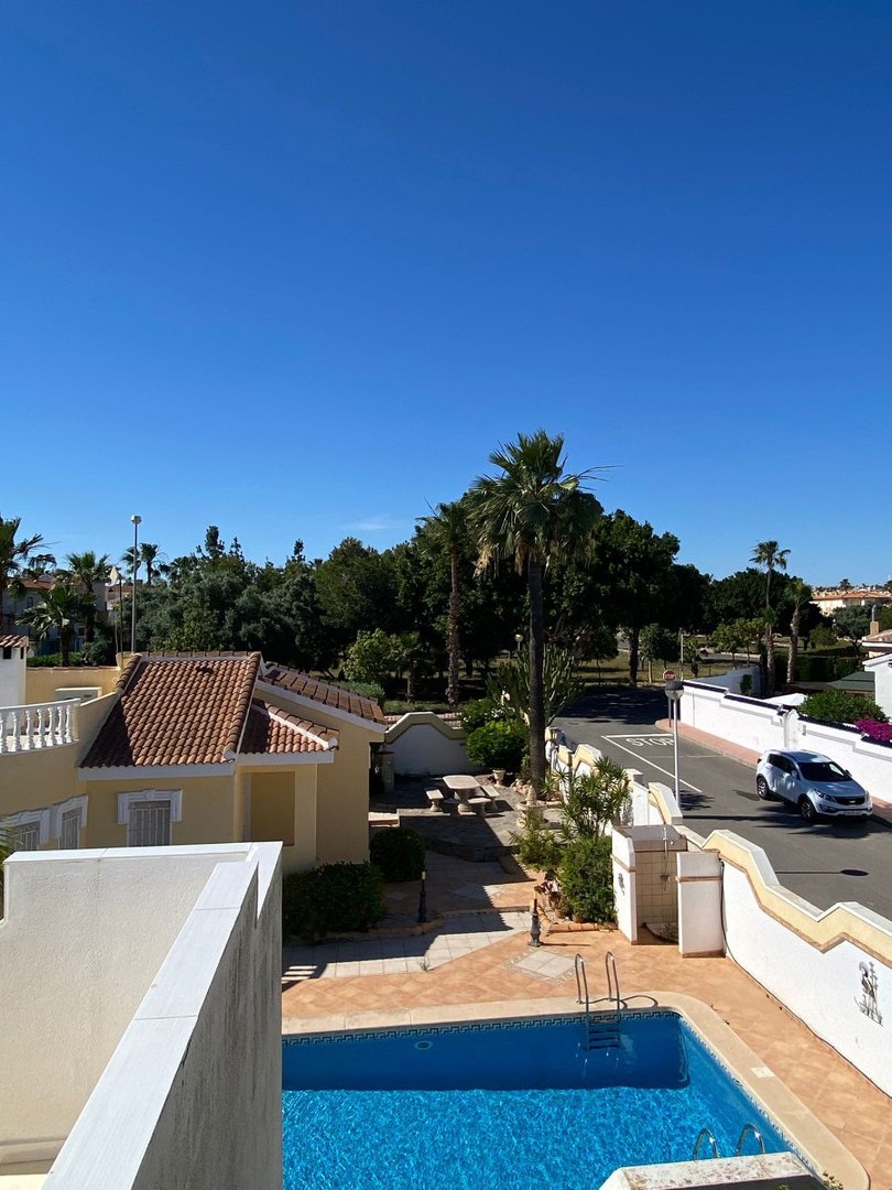 View from above a bright blue swimming pool and tiled patio, with a villa and palm trees under a clear blue sky.