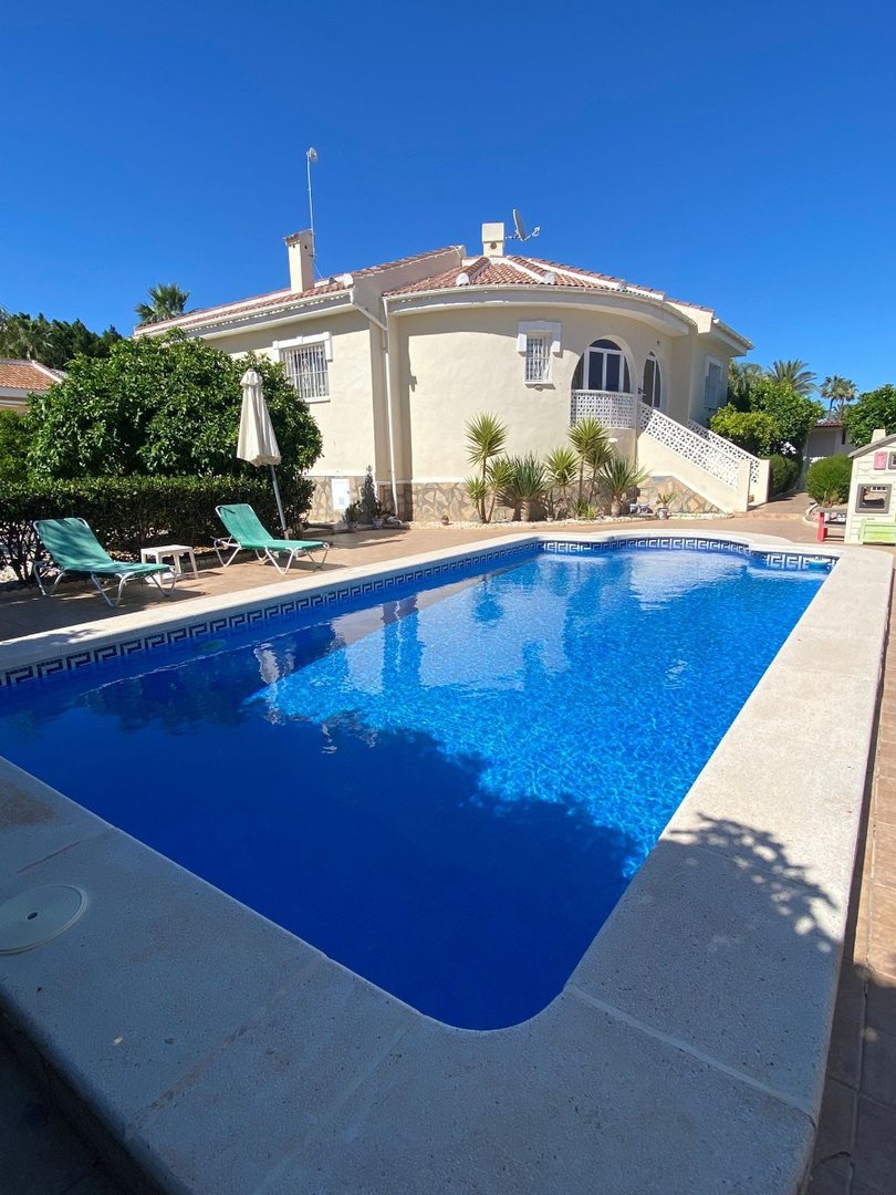 A beautiful villa with a large blue swimming pool, sun loungers, and lush greenery under a clear blue sky.