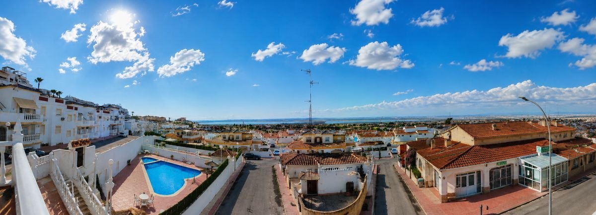 Panoramic view of a villa complex with a swimming pool and distant sea under a bright blue sky with clouds.