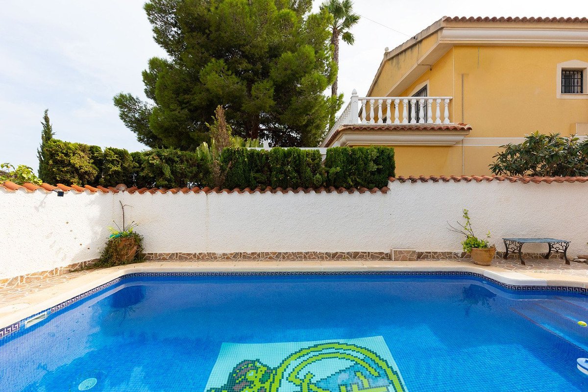 A vibrant blue swimming pool with mosaic art at the bottom, set against a backdrop of a white walled garden and a yellow villa.