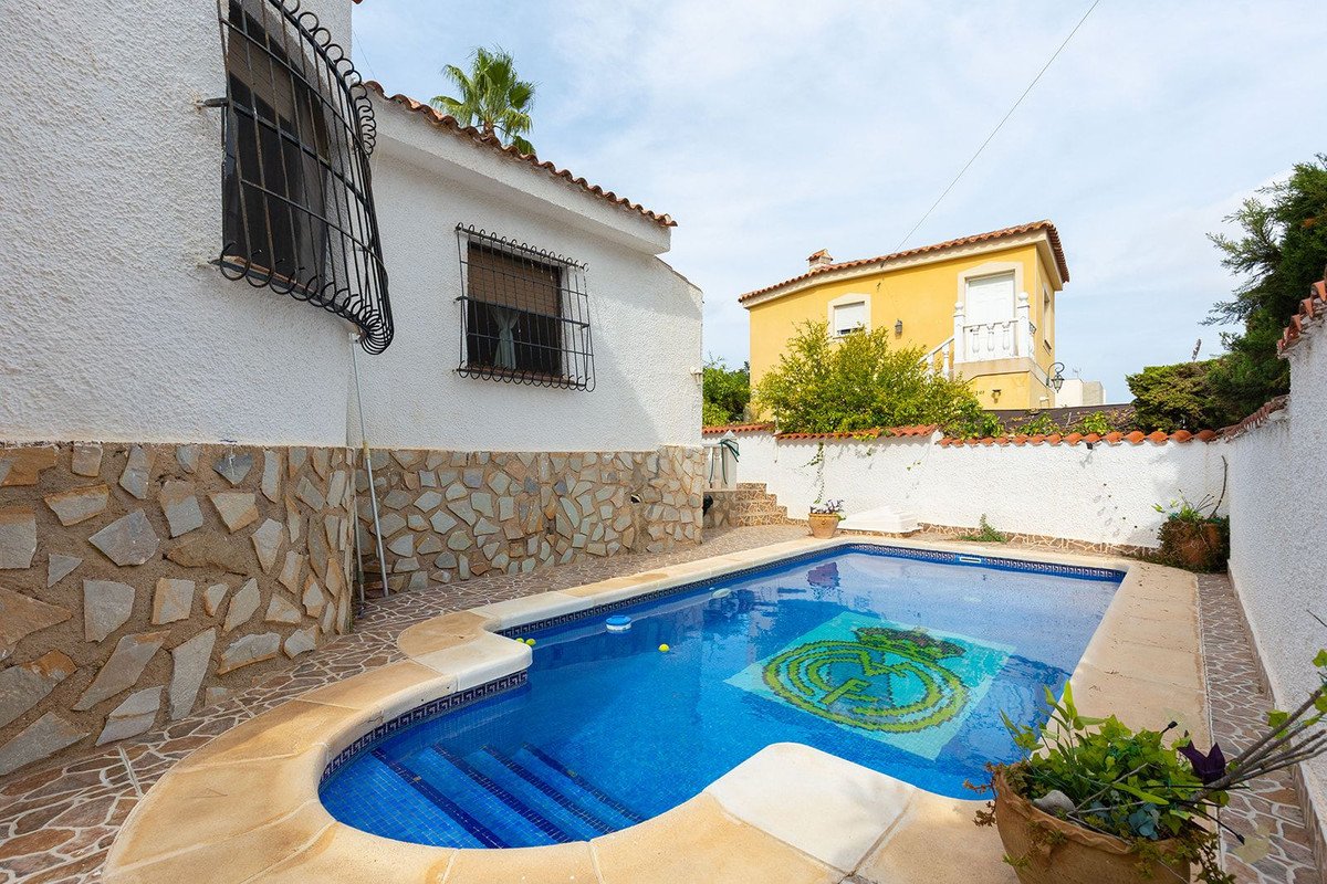 A bright blue swimming pool with a mosaic tile design at the bottom, surrounded by a paved patio and a white-stone-stone-clad house.