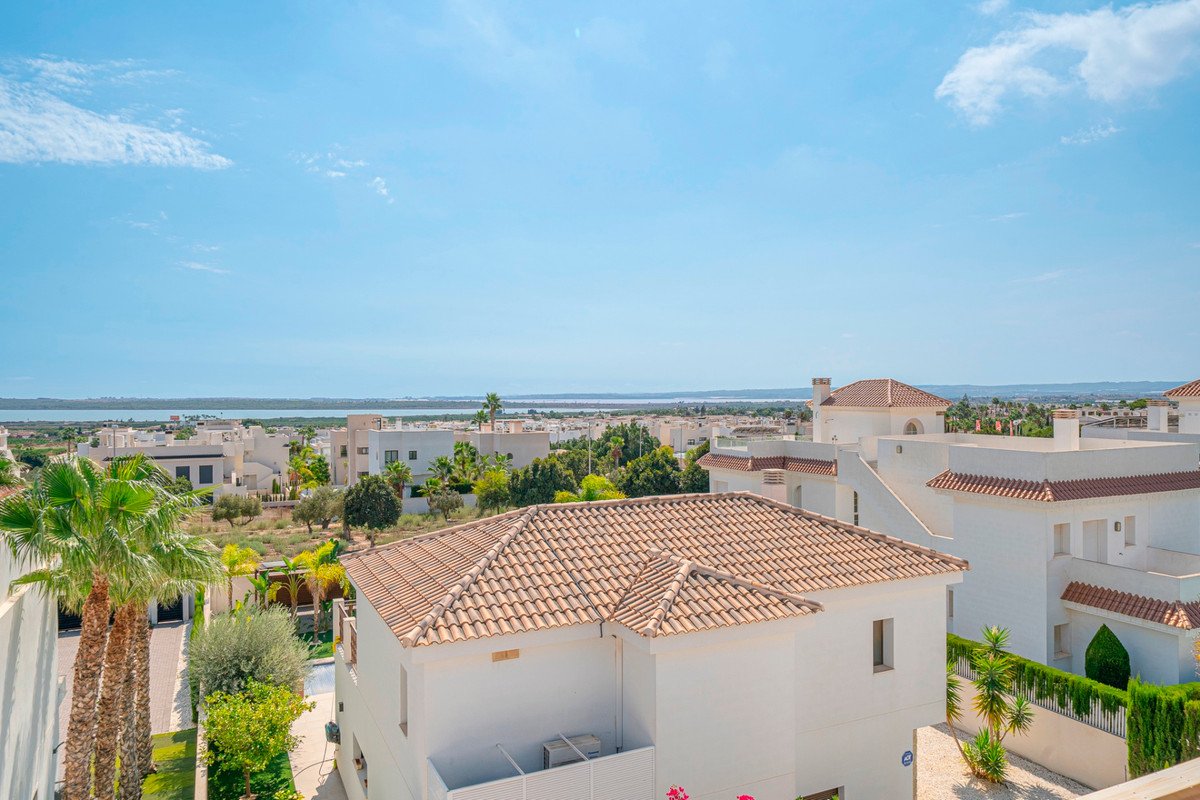 A wide shot shows a residential area with white buildings and tiled roofs, overlooking a tranquil sea under a blue sky.