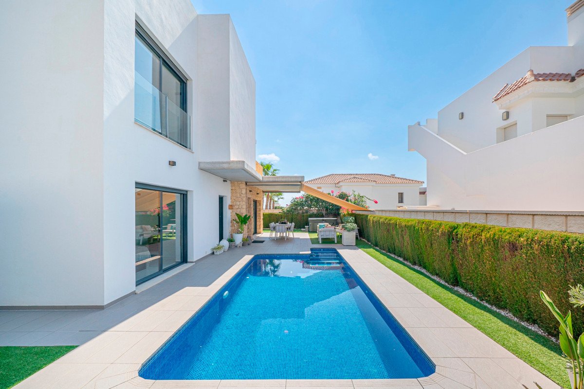 Modern white villa with a private swimming pool and green lawn, outdoor dining area, and sun loungers under a bright blue sky.