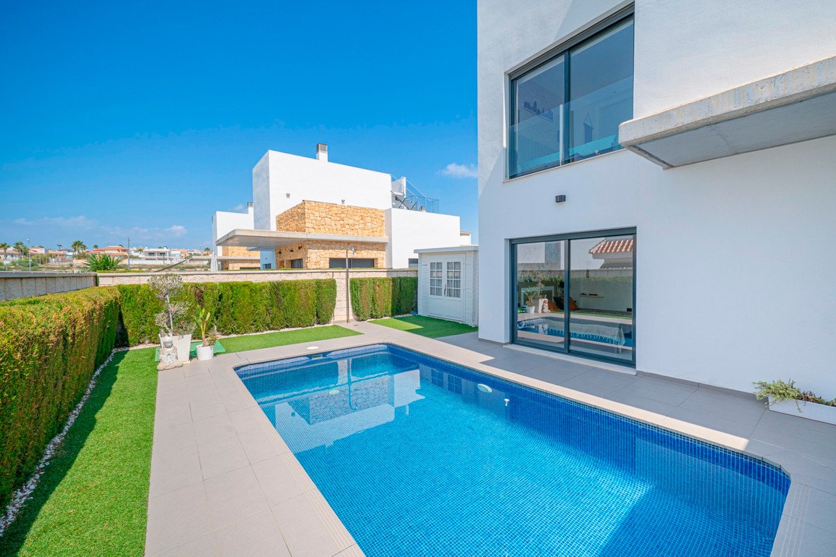 Modern villa exterior with a swimming pool, artificial grass, and manicured hedges under a clear blue sky.