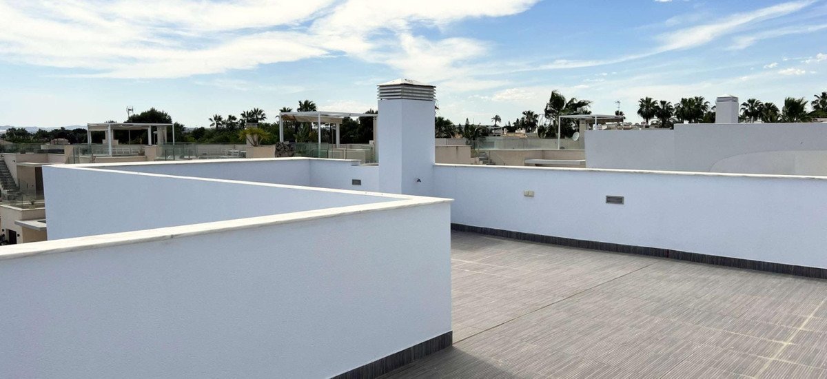 Modern terrace with white walls and tiled floor, overlooking palm trees and surrounding buildings under a cloudy sky.