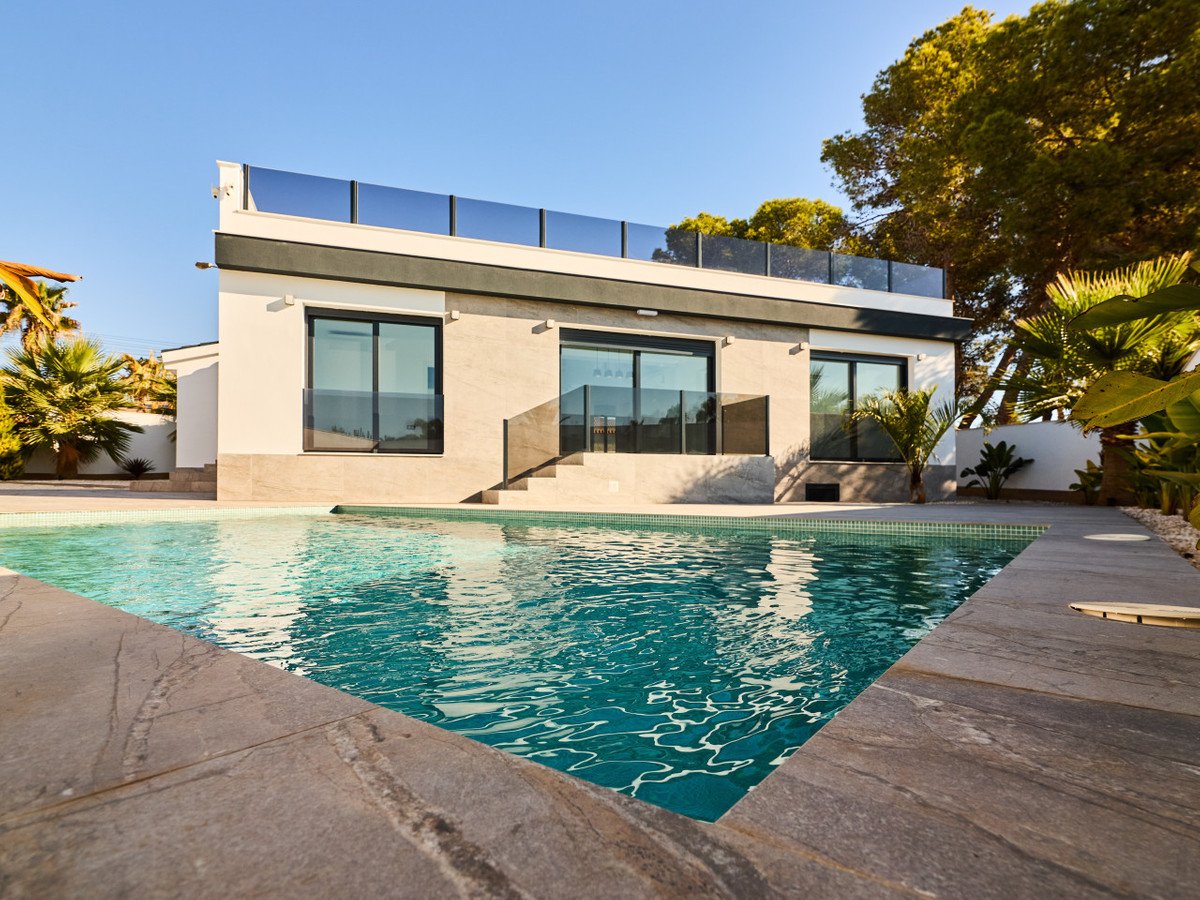 Modern white villa with large windows and a swimming pool in a sunny garden with palm trees.