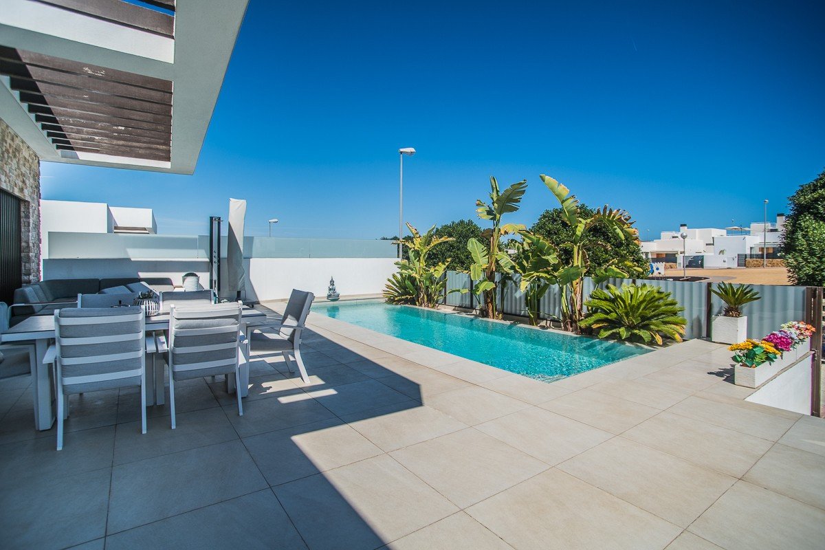 Modern villa with a swimming pool, outdoor dining area, lounge seating, and tropical plants under a clear blue sky.