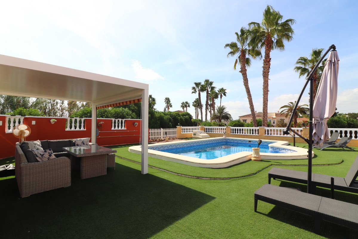 Outdoor patio with seating area, swimming pool, sun loungers, and palm trees under a clear sky.