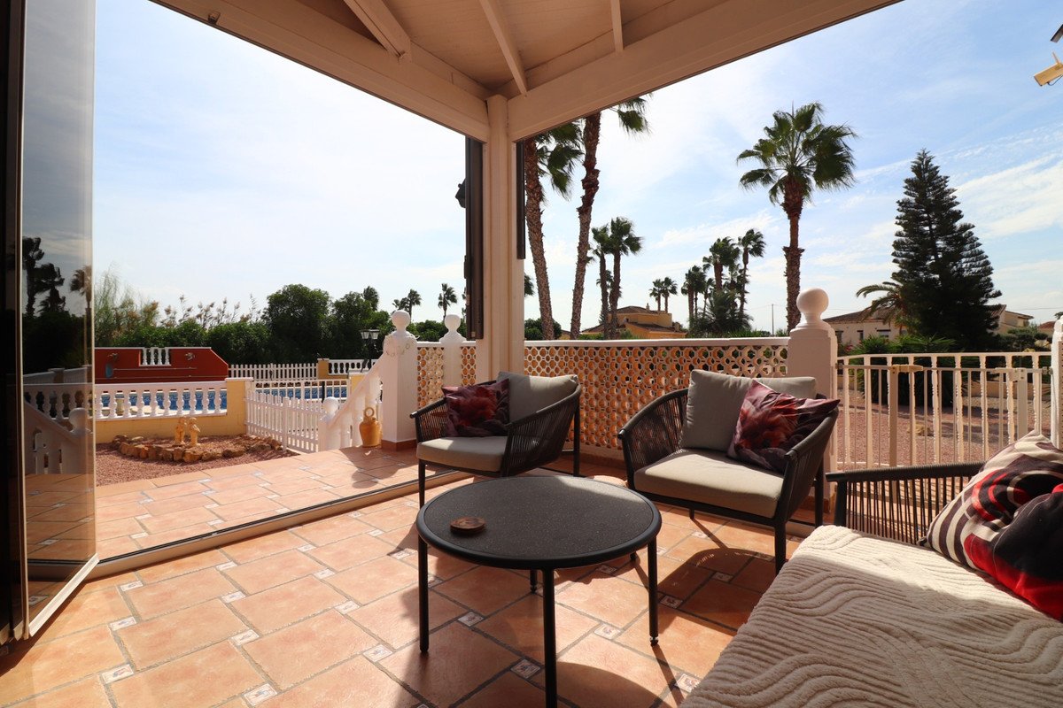 Covered terrace with seating and a table, overlooking a pool and palm trees under a clear blue sky.
