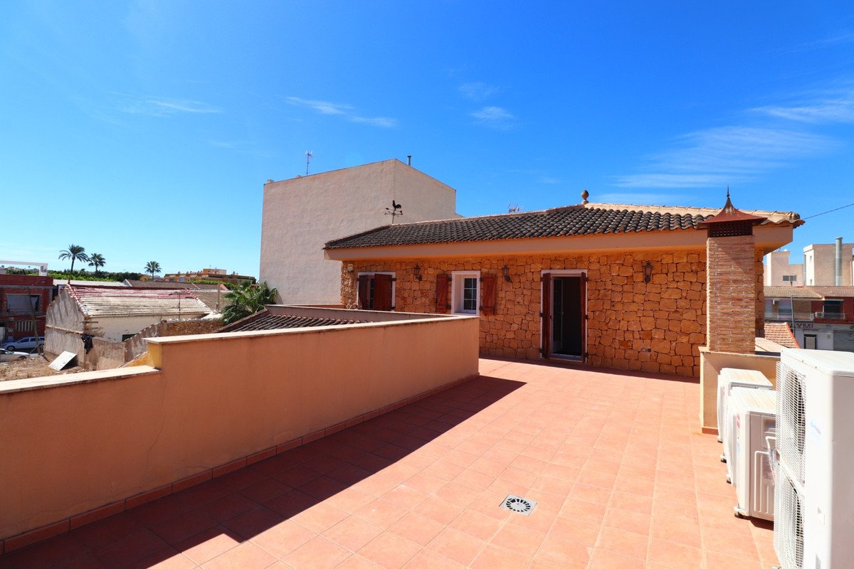 Terrace overlooking buildings with a stone-clad house featuring shutters and an outdoor chimney under a clear blue sky.