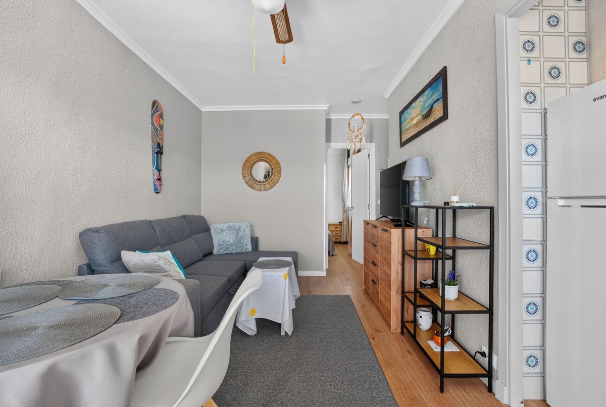 A compact living area featuring a grey sectional sofa, dining table, television, and decorative wall art, with a doorway leading to another room.