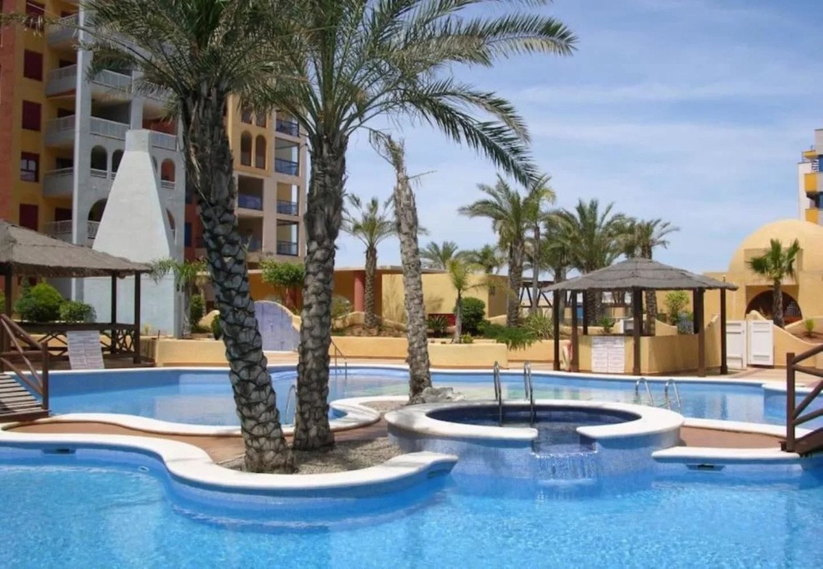 A resort complex with multiple swimming pools and palm trees, featuring a jacuzzi and covered cabanas.