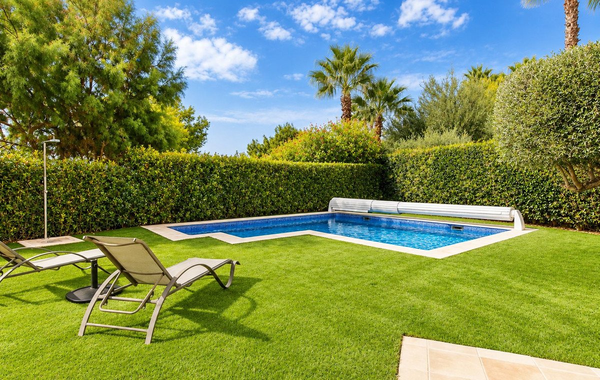 Outdoor swimming pool with a retractable cover, surrounded by green lawn, manicured hedges, palm trees, and lounge chairs on a sunny day.