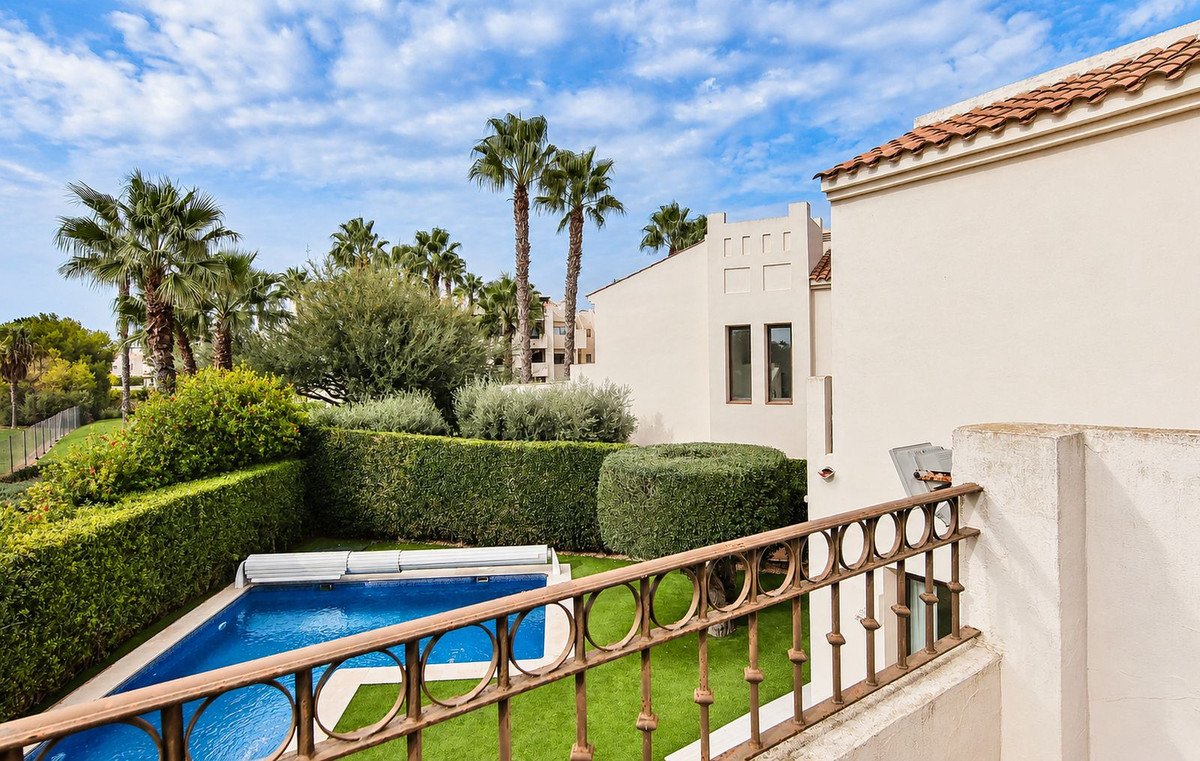 View of a private swimming pool and lush garden with palm trees and manicured hedges, from a balcony with a decorative railing.