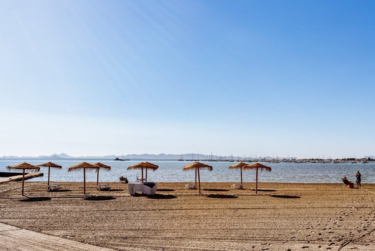 Beach with thatched umbrellas and lounge chairs overlooking a calm sea with a distant marina and mountains under a clear blue sky.