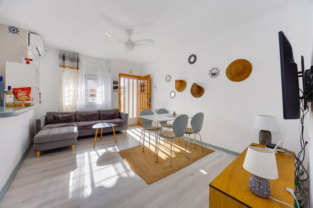 Bright living and dining area with a grey sofa, dining table with chairs, and a mounted television, featuring natural light.