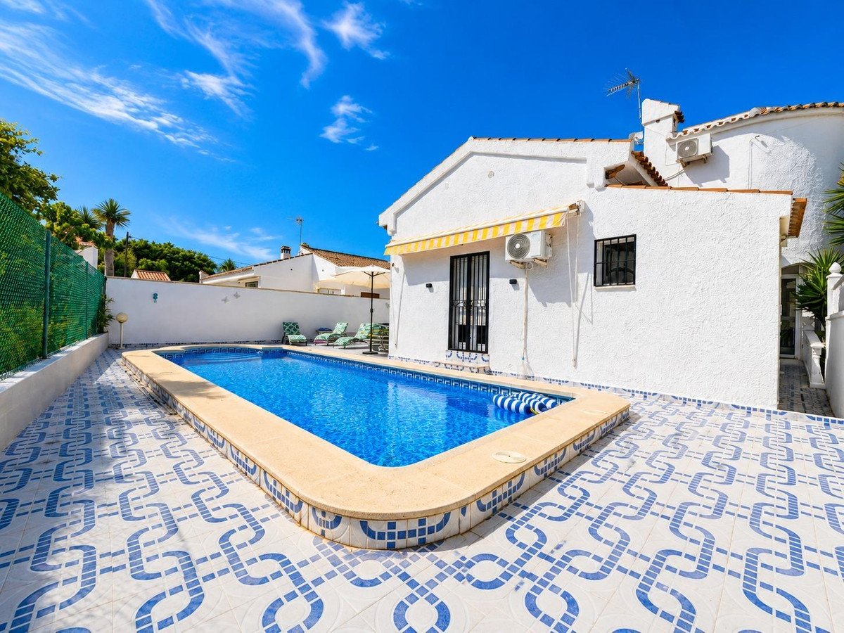 Exterior view of a villa with a private swimming pool, sun loungers, and a patterned tiled patio on a sunny day.