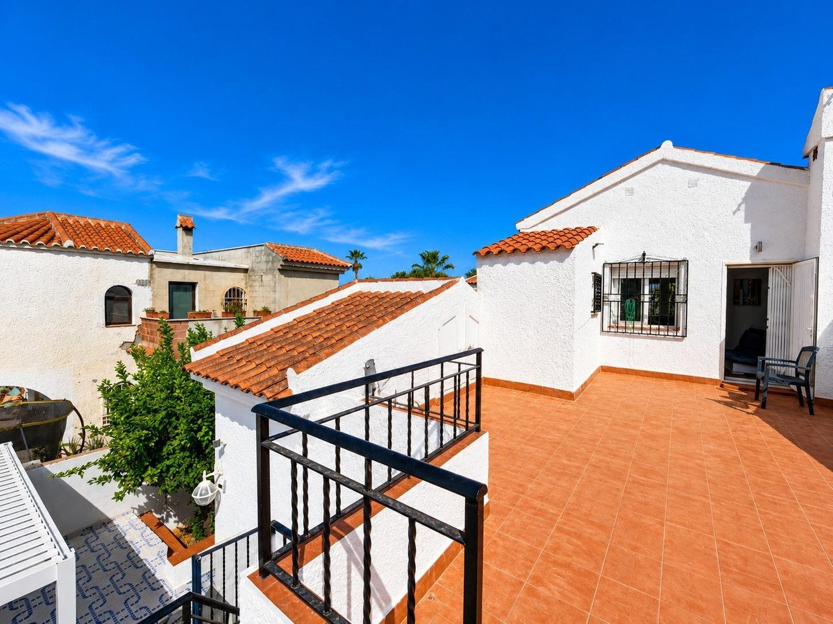 A spacious terrace with terracotta tiles and a black metal railing overlooks several Spanish-style villas under a clear blue sky.