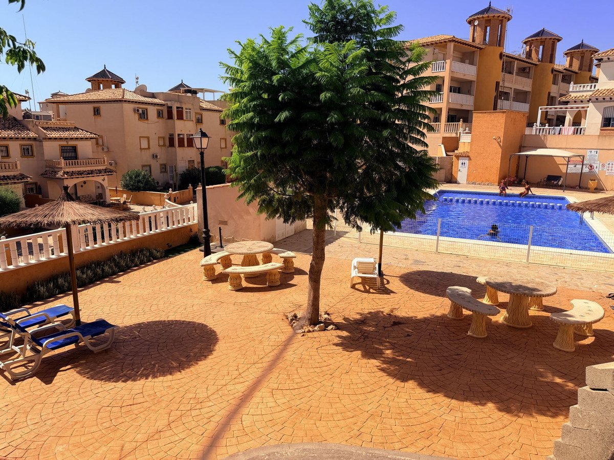 A sunny resort courtyard with a swimming pool, picnic tables, lounge chairs, and surrounding buildings.