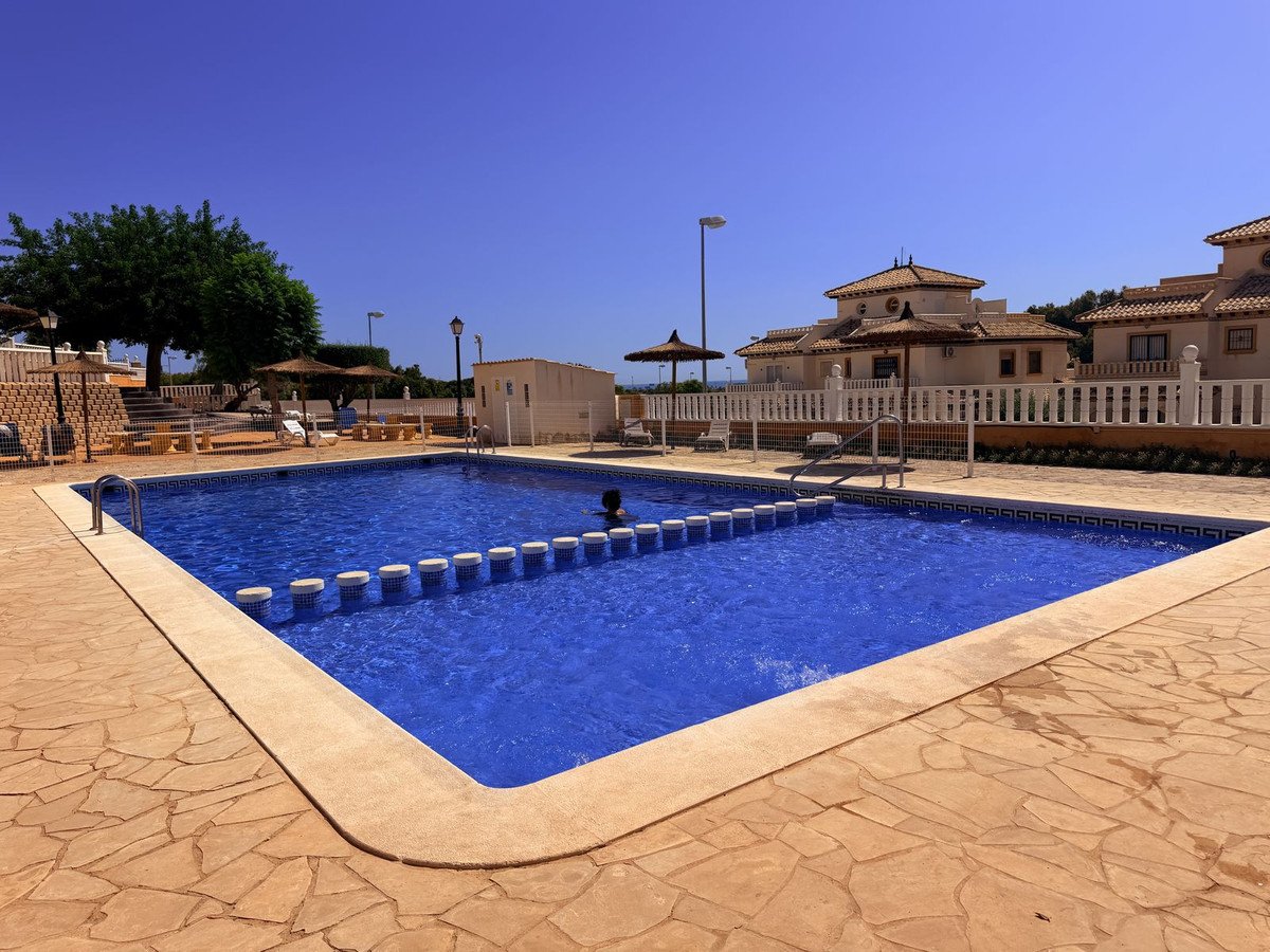 A large blue swimming pool with a paved deck, surrounded by fencing and houses, under a clear blue sky.