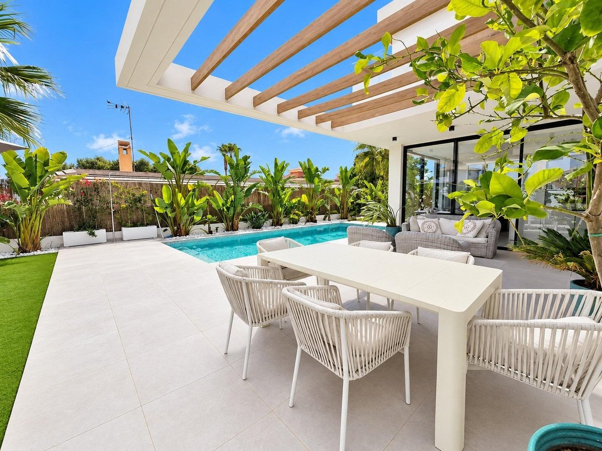 Modern patio with outdoor dining table and chairs next to a swimming pool and lush tropical plants under a pergola.