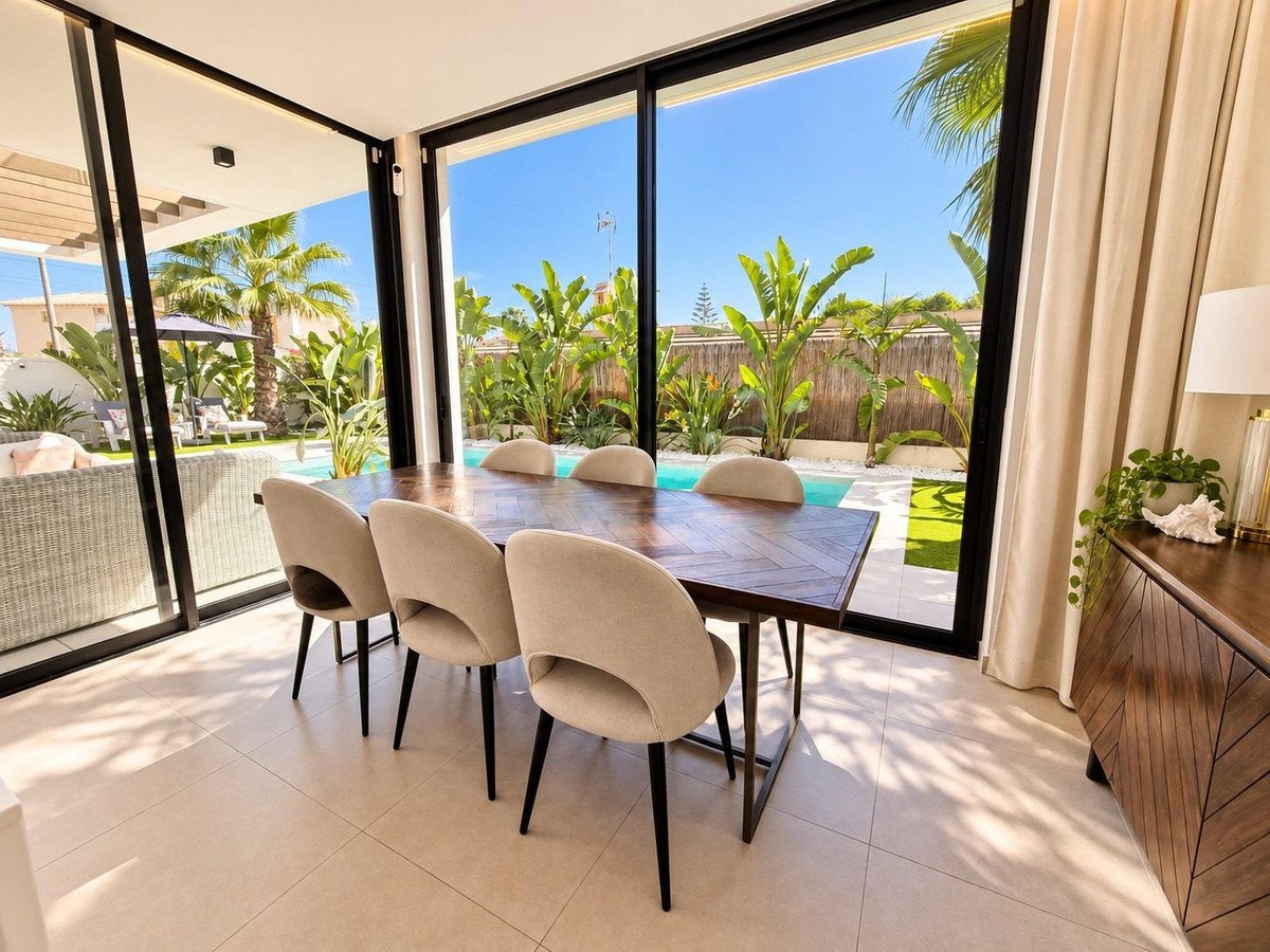 Modern dining area with pool and garden view, featuring a large wooden table and comfortable chairs, with sliding glass doors leading outside.