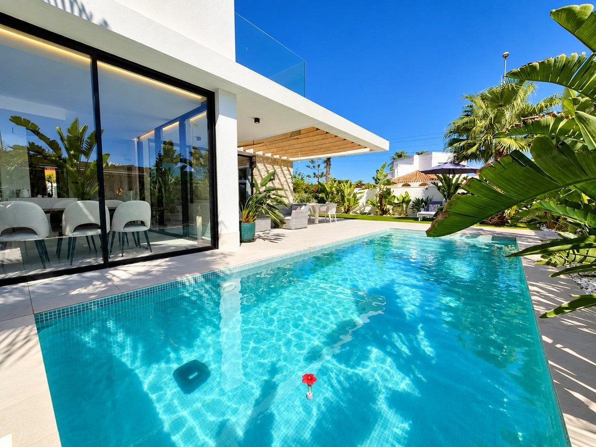 Modern villa with a swimming pool, outdoor dining area, and a glimpse of the interior dining room through large glass doors.