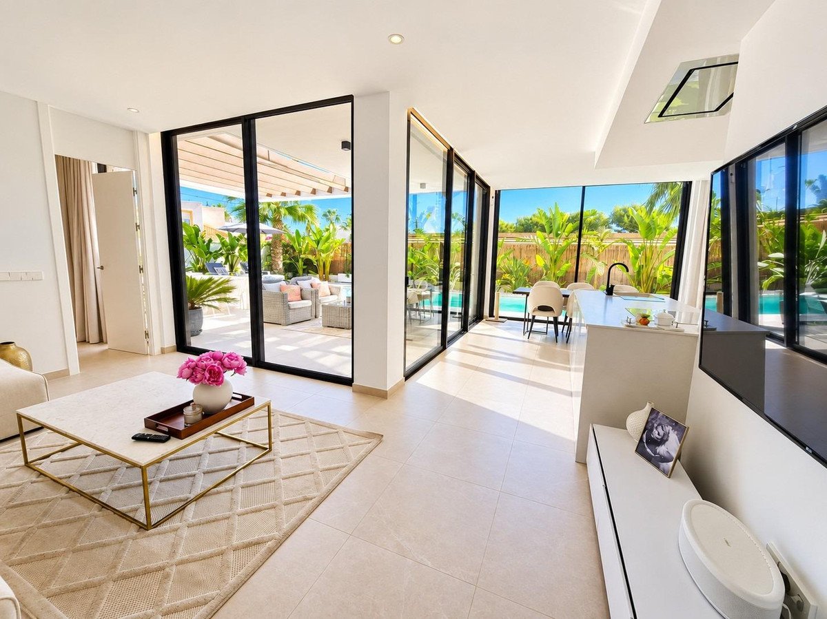 Modern living room with open-plan kitchen, featuring large glass doors opening to a patio and pool with lush tropical landscaping.