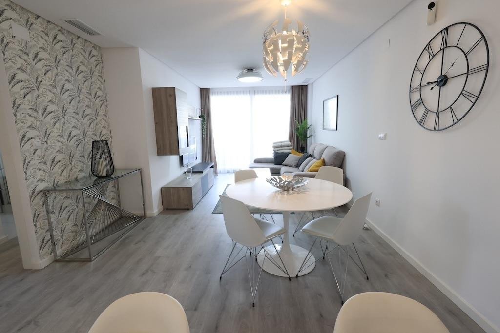 Modern living and dining area with sectional sofa, round table, white chairs, and large wall clock. Bright natural light.