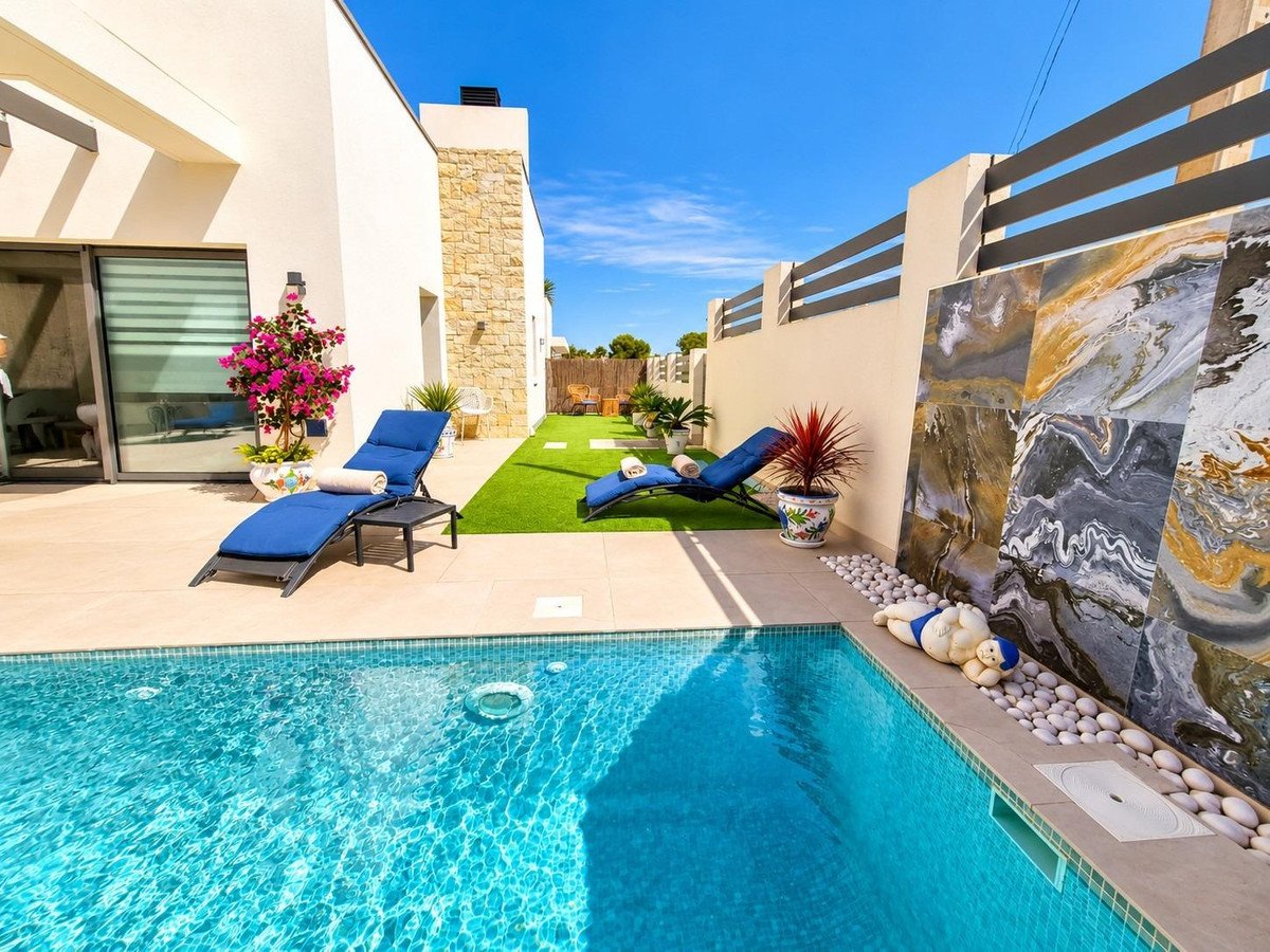 Modern villa with a sparkling swimming pool, sun loungers, artificial turf, and decorative wall tiles under a clear blue sky.