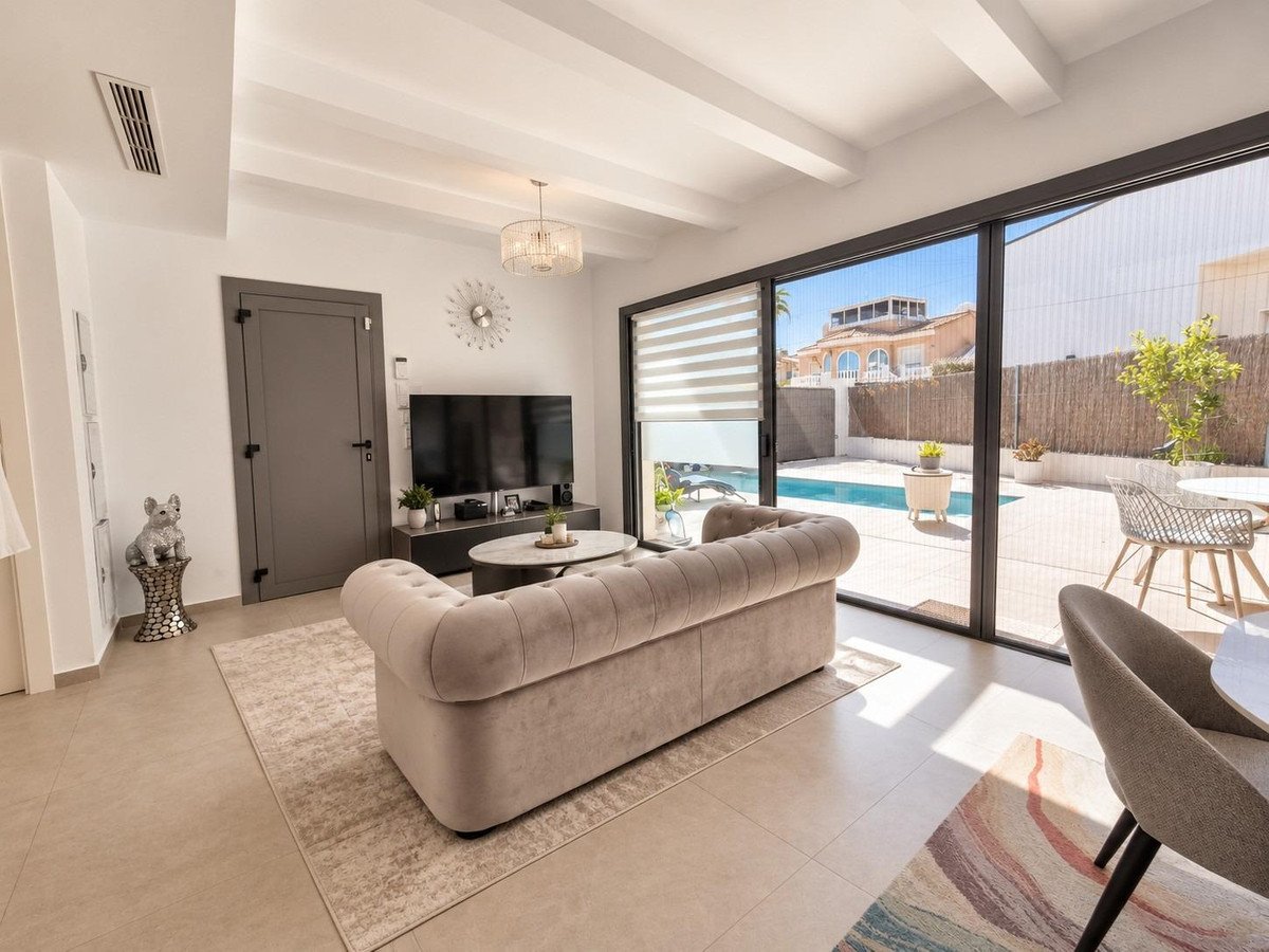 Modern living room with a beige chesterfield sofa, television, and sliding doors opening to a private pool and patio area.