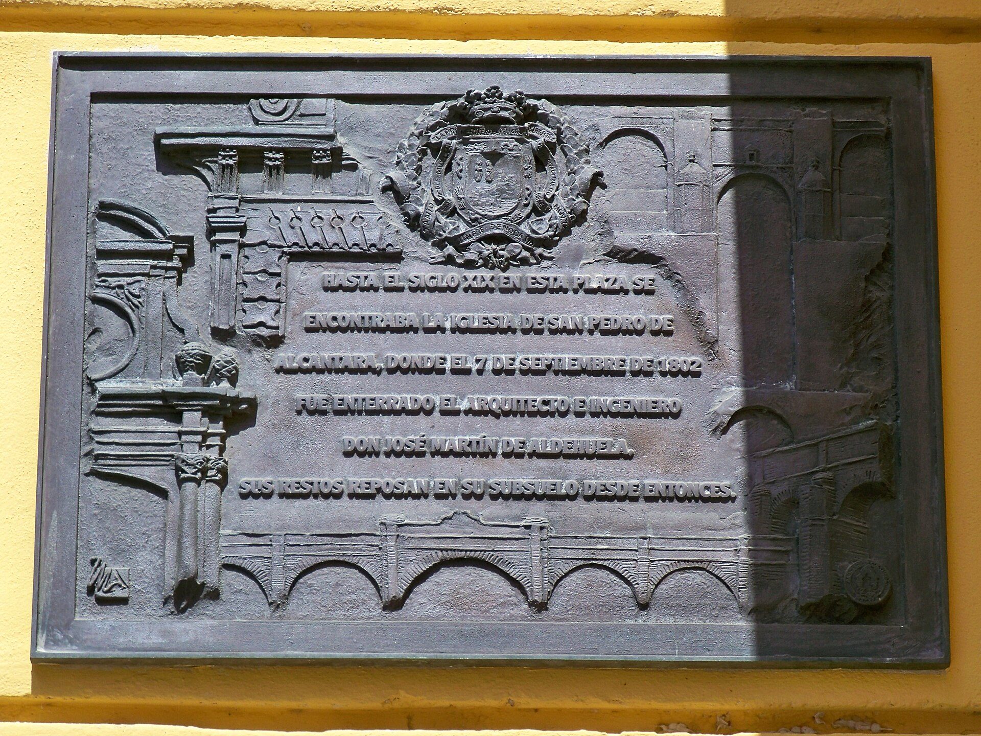Alt text: Plaque with historical text and coat of arms on a yellow wall.