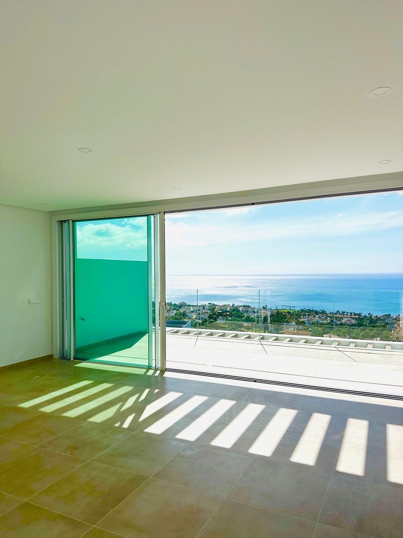Empty bedroom with sliding glass doors opening to a terrace with a stunning sea view and distant coastal town.