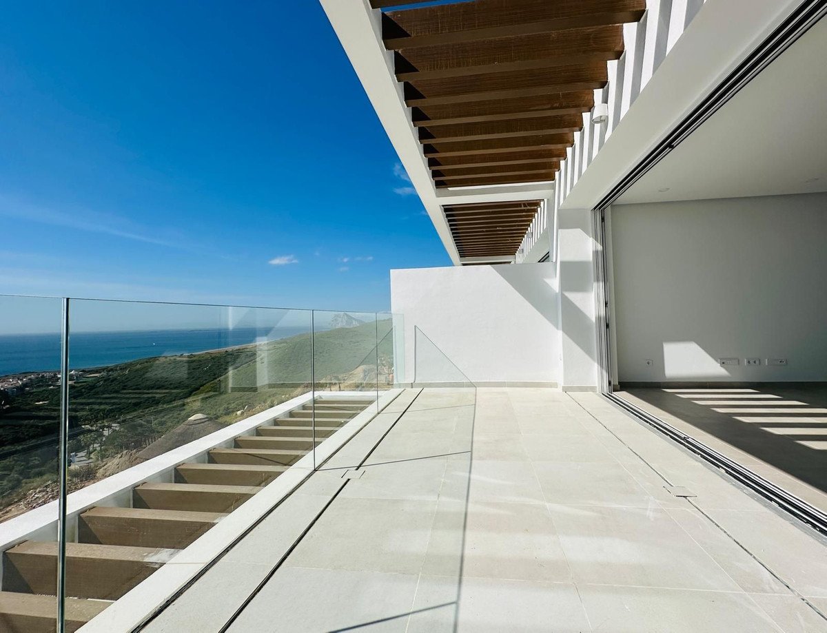 Modern terrace with glass railings overlooking the sea and a scenic hillside. Features a wooden pergola and large sliding doors.
