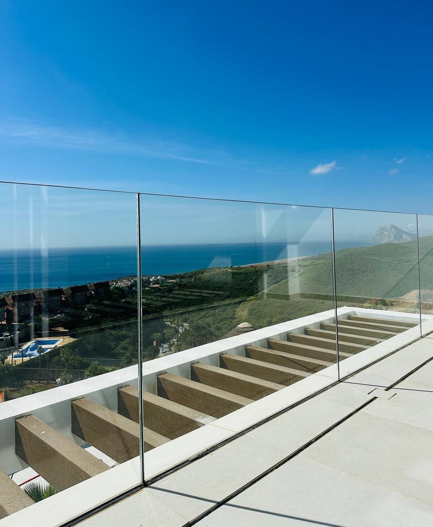 Modern terrace with glass railing offering panoramic sea and coastal views under a clear blue sky.