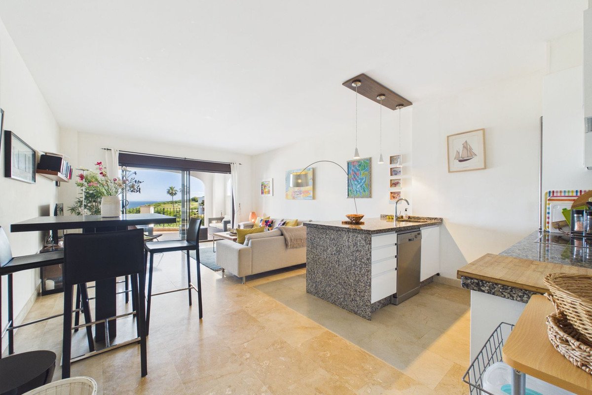 Open-plan living, dining, and kitchen area with a high bar, comfortable sofa, and a view of the sea and palm trees.