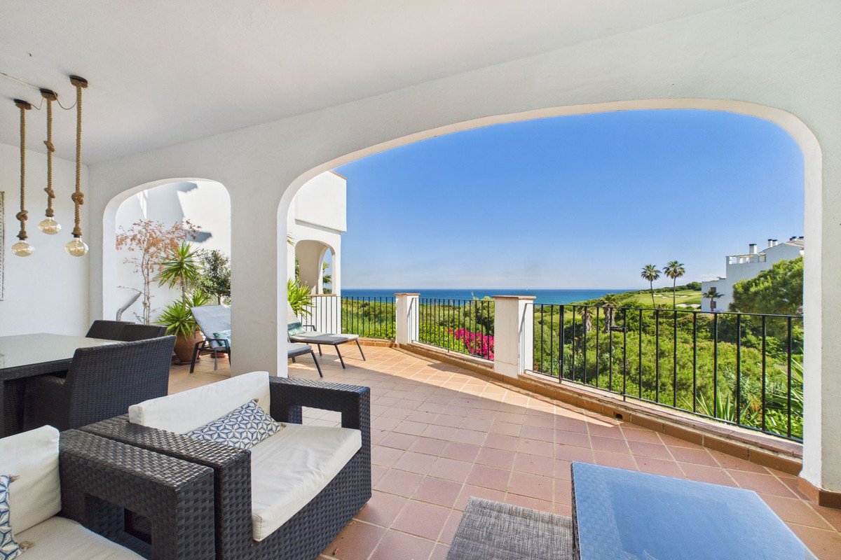 Covered terrace with outdoor seating overlooking a lush green golf course and the blue sea on a sunny day.