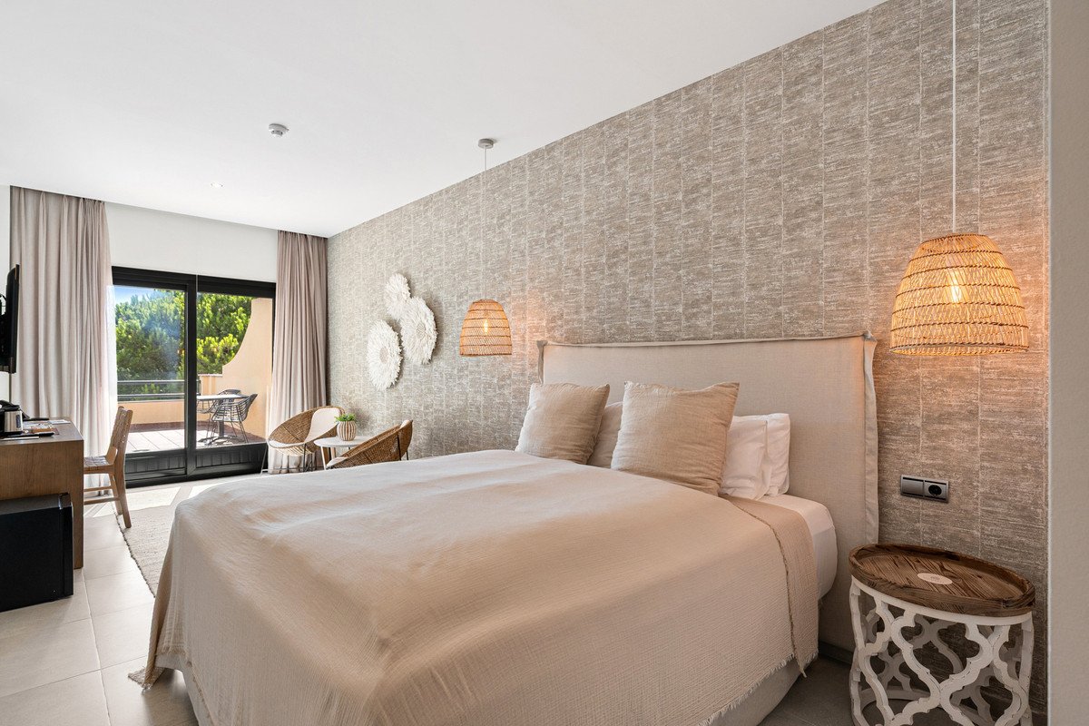 A beige bedroom with a king-size bed, textured wallpaper, pendant lights, and a balcony with trees visible.