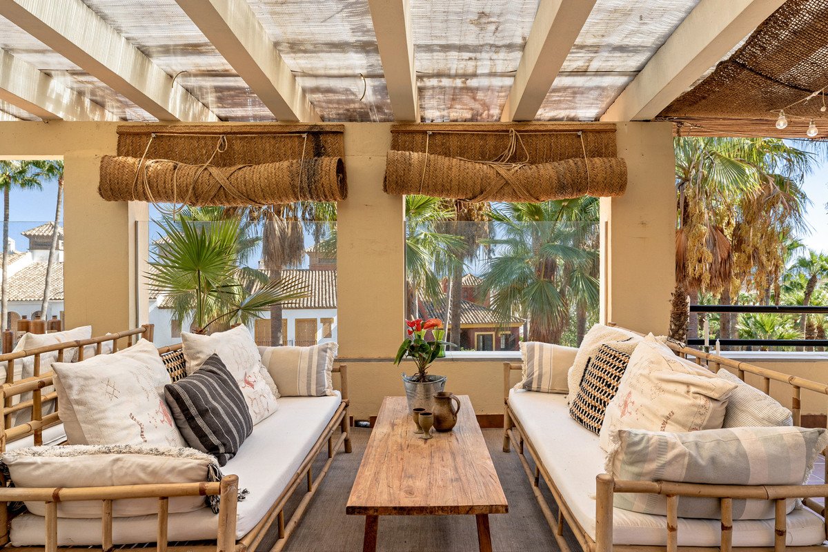 A covered patio with bamboo sofas, a wooden coffee table, and views of palm trees and Spanish architecture.