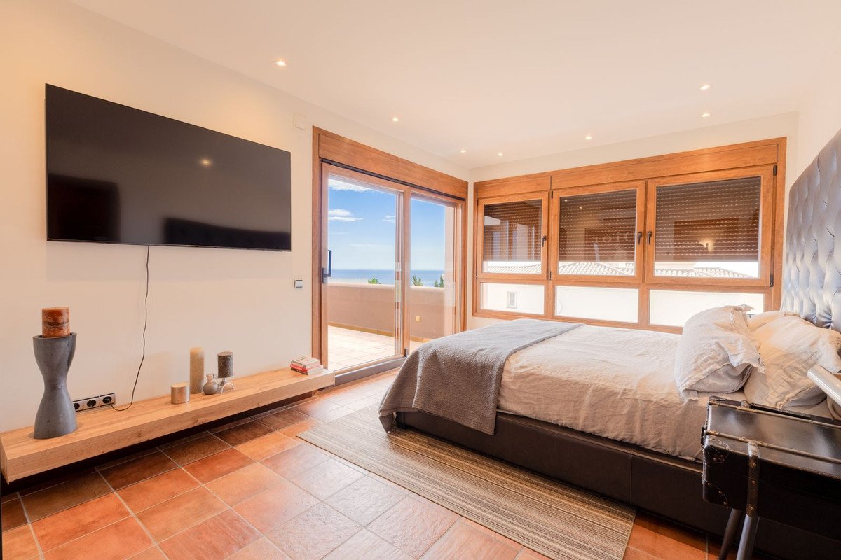Bedroom with a large bed, a wall-mounted TV, sliding doors opening to a terrace with a sea view, and wooden window frames.