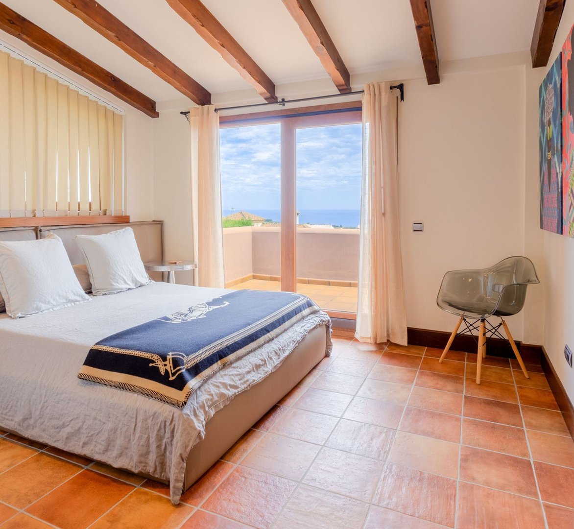 Bedroom with a large bed, wooden beams on the ceiling, and a balcony with a stunning sea view.